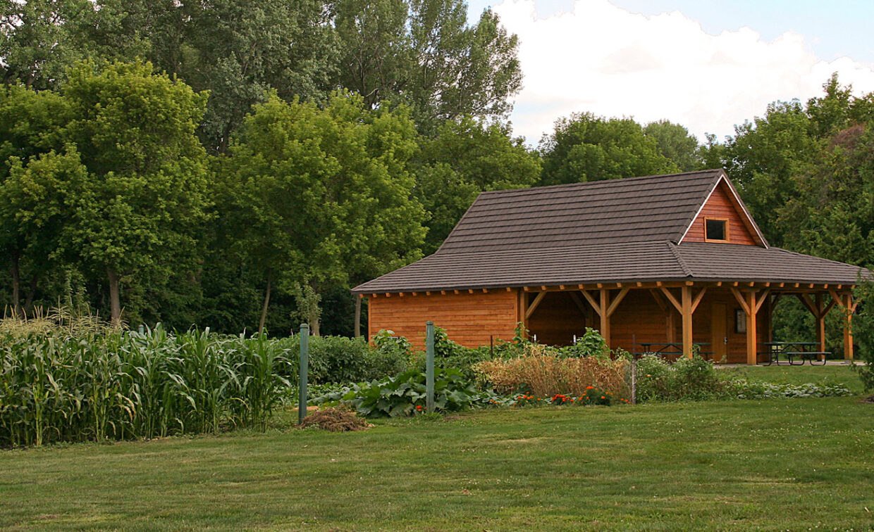 A brown wooden pavilion with a dark roof stands among tall green trees and a community garden with rows of corn and other plants.