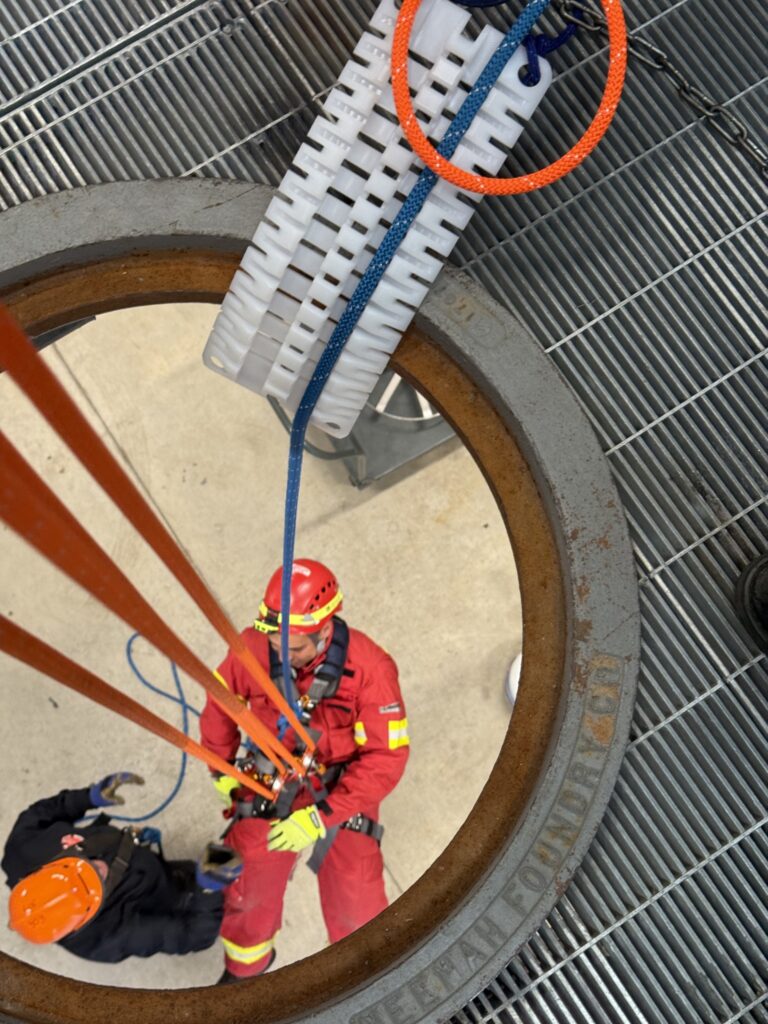 Query successful
A firefighter in red protective gear and a helmet descends into a confined space with ropes, while another firefighter in black and an orange helmet lies on the ground below.