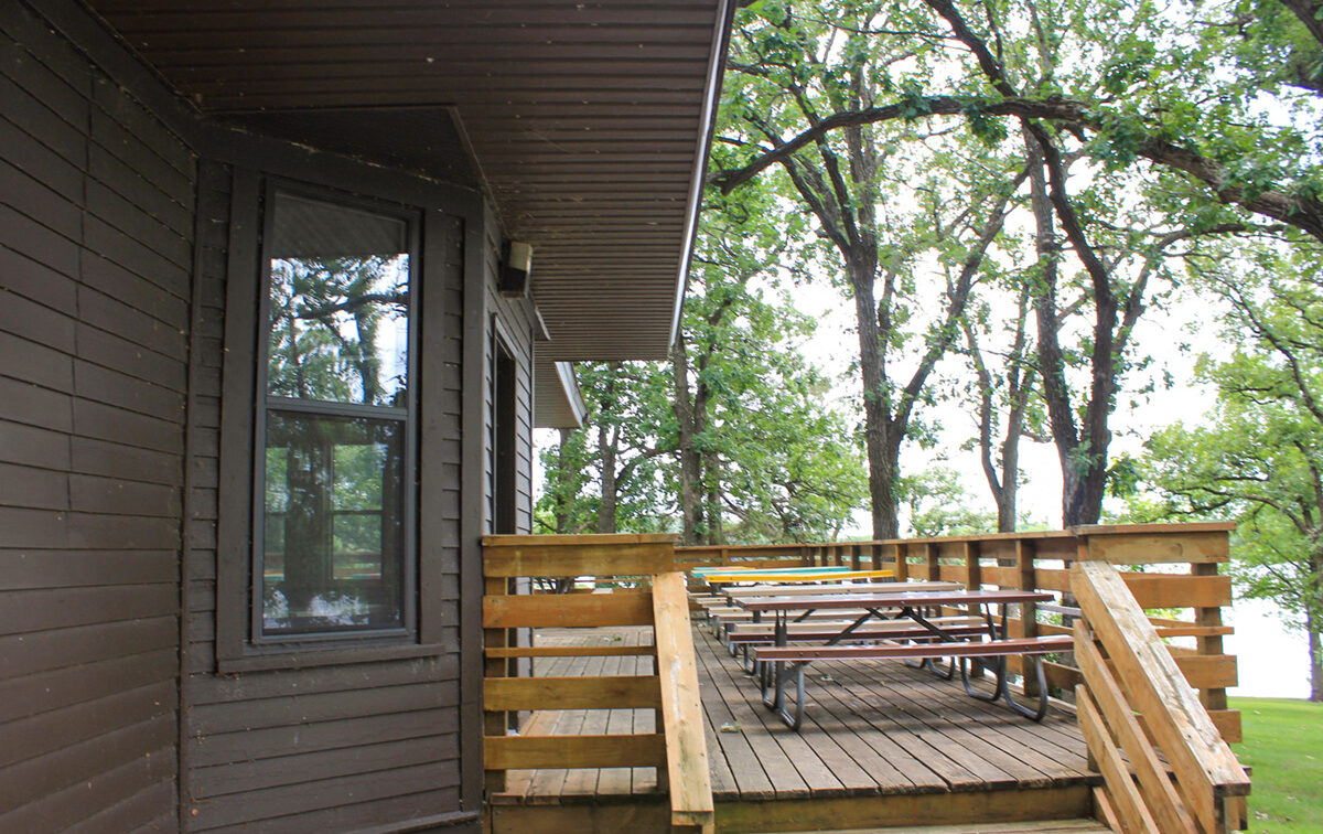 A wooden deck of a brown cottage, featuring picnic tables, with tall trees visible in the background under an overcast sky.