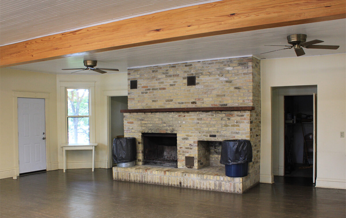 Large, light-colored brick fireplace with two fireboxes and a wooden mantel, flanked by two trash cans, in a room with a wood-beamed ceiling.