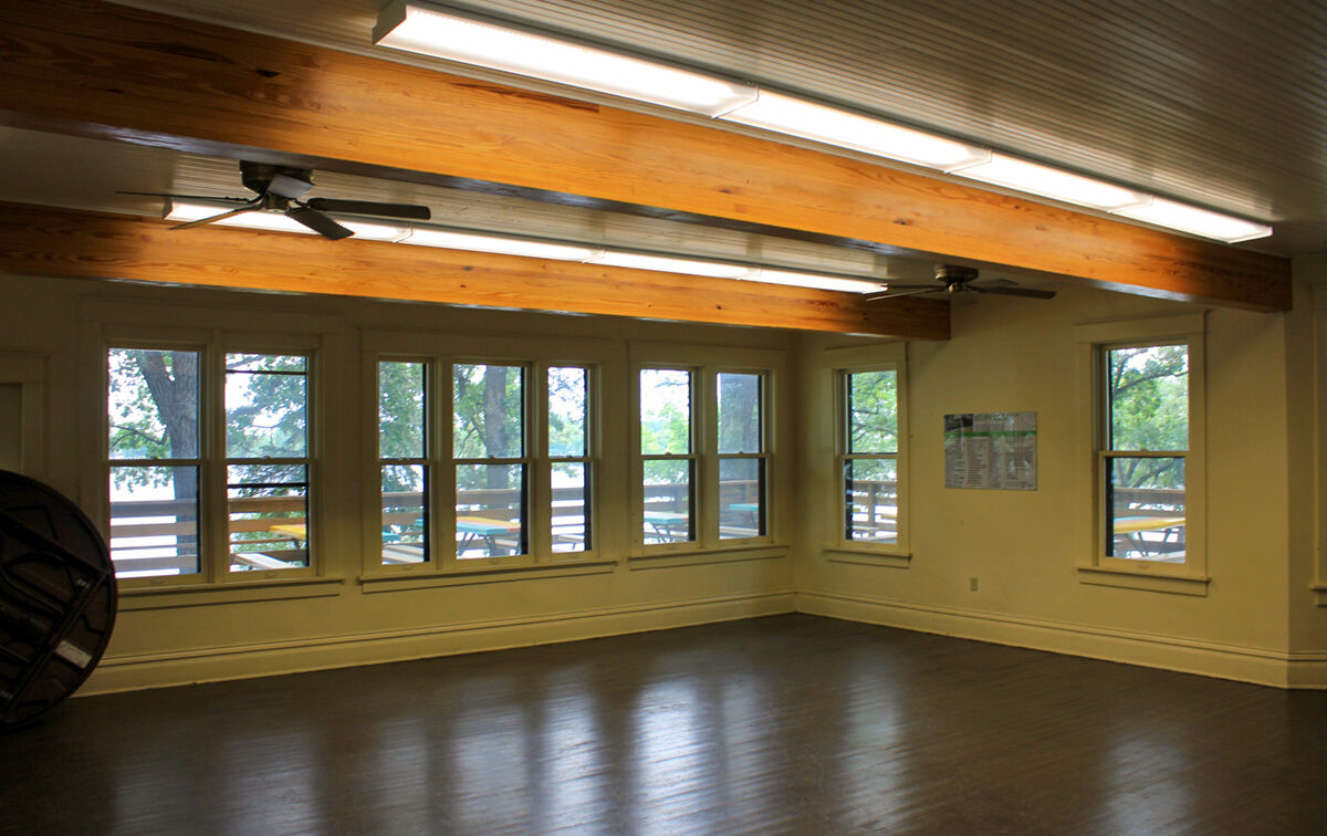 Interior of a cottage room with dark wooden floors, light walls, and several large windows offering a view of an outdoor deck and trees. The ceiling has wooden beams, fluorescent lights, and ceiling fans.