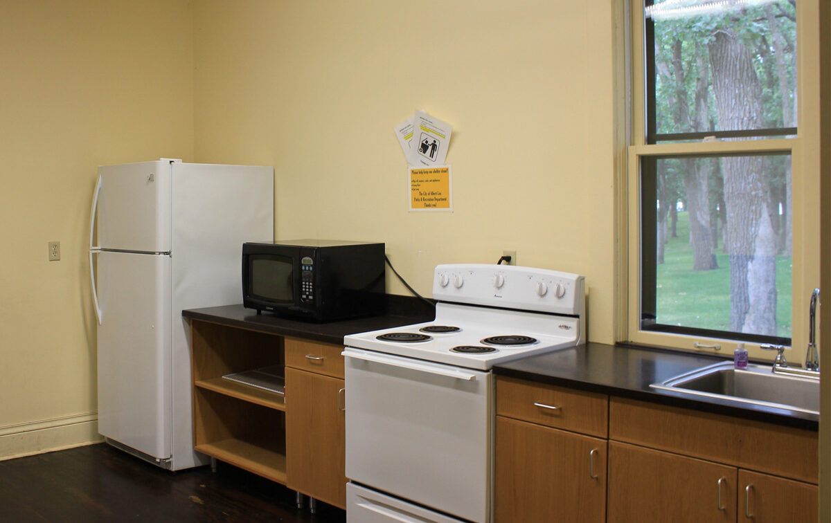 A simple kitchen in Albert Lea, Minnesota, with a white refrigerator, a black microwave on a dark countertop, a white electric stove, and light wooden cabinets against a pale yellow wall.