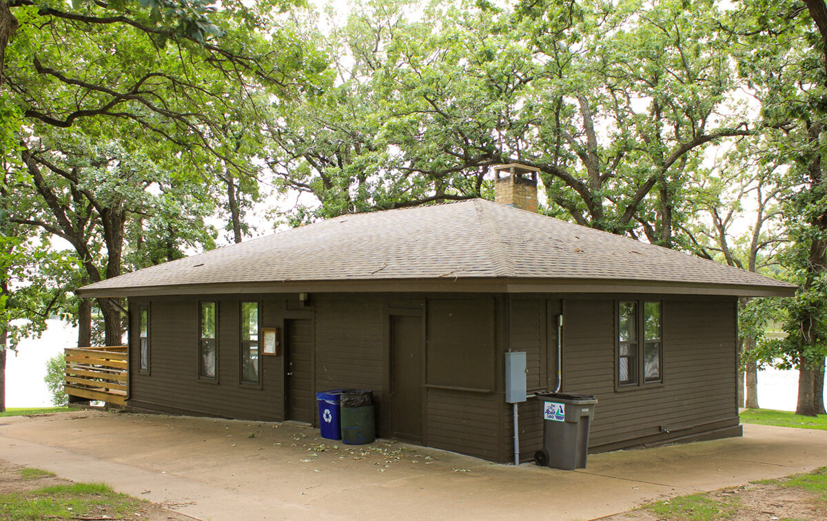 Exterior of a dark brown, octagonal cottage with a brown roof and a brick chimney, surrounded by lush green trees, with a recycling bin and trash can outside.
