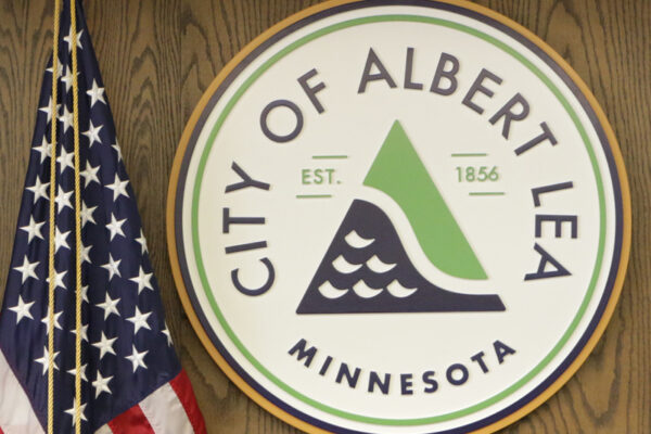 The City of Albert Lea, Minnesota logo, featuring a green and blue triangular design, next to a folded American flag.