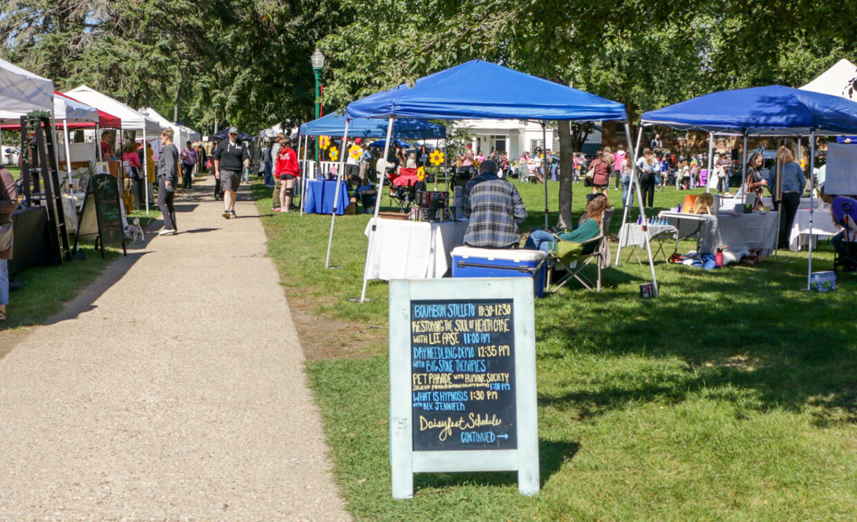A wide outdoor shot shows a bustling daytime market scene under a bright sky. A light-colored dirt path runs from the foreground into the middle distance on the left, lined by several white pop-up tents with people Browse. On the right, more tents, some blue, are set up on a grassy area, with visitors mingling and vendors tending to their stalls.