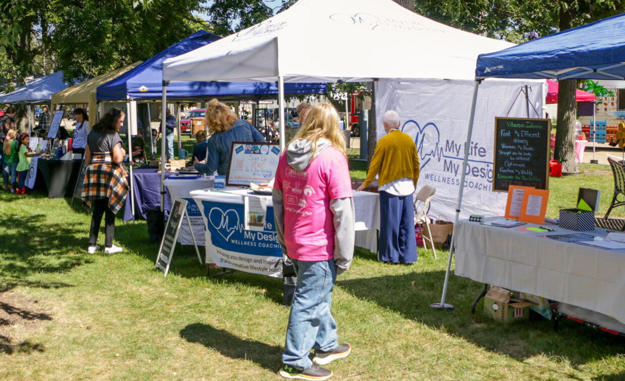 A slightly low-angle, wide shot captures a lively outdoor market scene. Several white and blue tents are set up on a vibrant green lawn, under dappled sunlight. In the foreground, a person in a bright pink hoodie and jeans stands with their back to the viewer, looking towards a white tent. Under this tent, a "My Life My Design Wellness Coaching" banner is visible on a table, with people interacting around the stall.