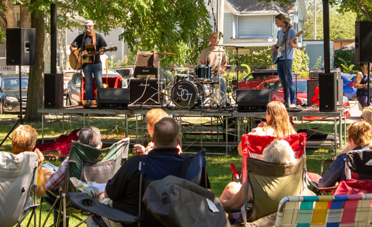 A sunny outdoor scene shows a band performing on a raised stage in a park. Three musicians are visible: a guitarist on the left playing an acoustic guitar, a drummer in the center behind a drum kit with "507" on the bass drum, and another guitarist on the right playing an electric guitar.
