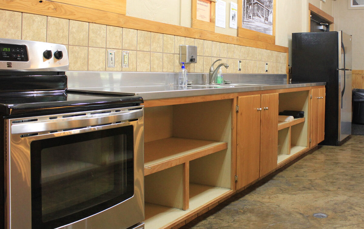 The image shows a kitchen with a stainless steel stove, a long stainless steel counter with a sink, and light wooden cabinets. The kitchen has a tiled backsplash, wooden trim, and a concrete floor. A black refrigerator is visible on the right.