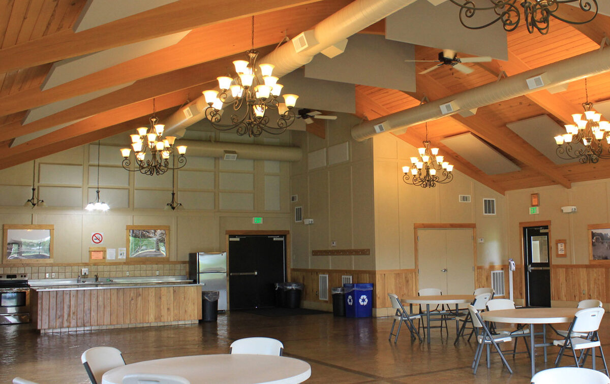 The image depicts the interior of a large hall with a high vaulted wooden ceiling and chandeliers. It includes a kitchen/serving area with a counter and sink, and multiple round tables with chairs set up in the main space.