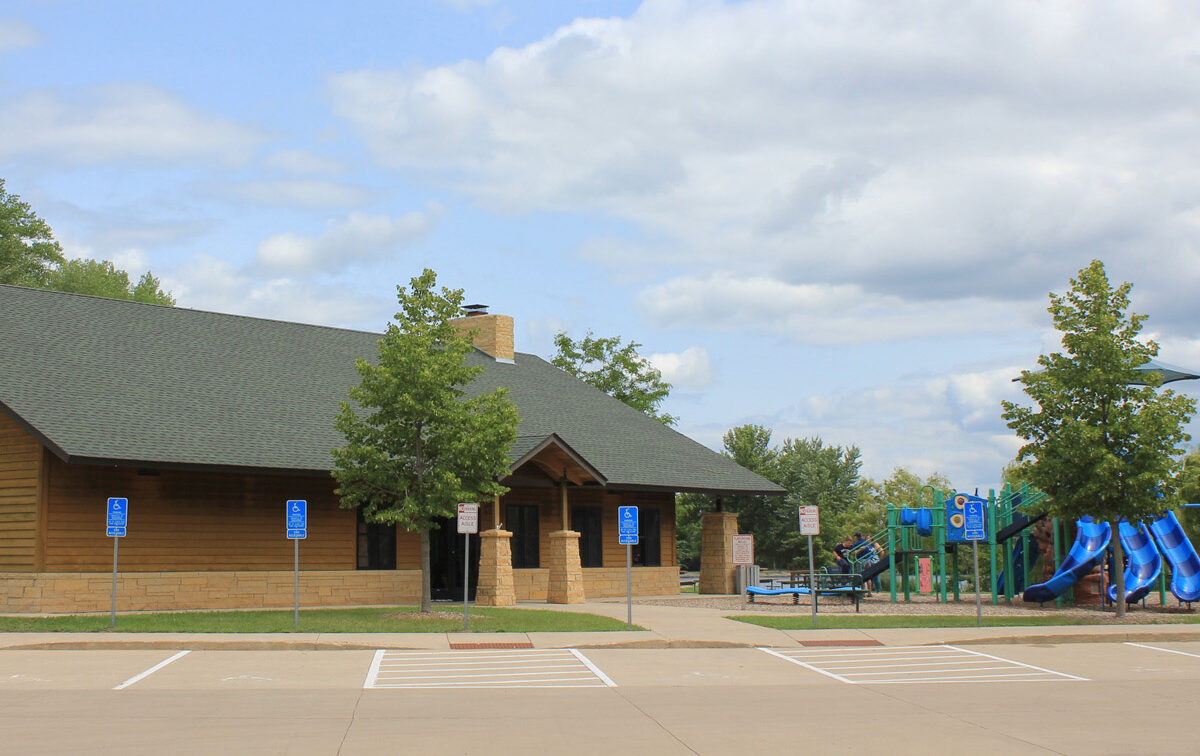 Exterior view of a park building with a green roof and wooden siding, next to a playground with blue slides, under a partly cloudy sky.