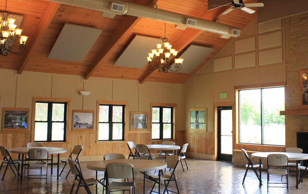Bright community hall with vaulted wooden ceiling, chandeliers, round tables, and chairs. Natural light fills the room through large windows.