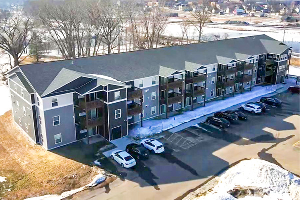 An aerial view of a long, modern apartment building with a dark gray roof and multi-toned gray siding, surrounded by a parking lot with cars, and bare trees in a winter landscape.