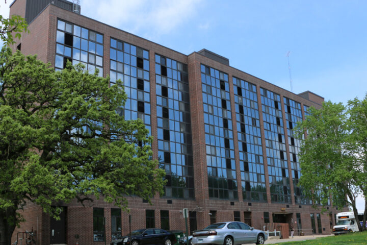 A large, multi-story brick building with many windows reflects the sky. Two cars are parked on a paved area in front of it. There are green trees on either side of the building, and a grassy lawn slopes down in the foreground. A white van is parked on the right side of the building, further back.