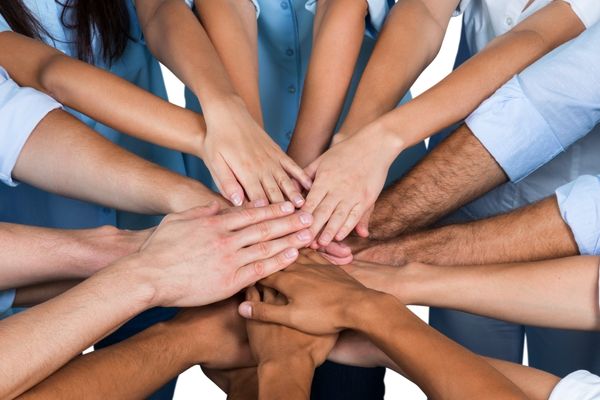 A high-angle close-up shot shows a diverse group of hands, belonging to approximately ten individuals, stacked on top of one another in a gesture of unity or teamwork. The hands vary in skin tone, ranging from light to dark, emphasizing diversity.