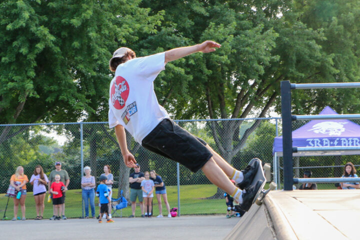 A low-angle, full shot shows a person on a skateboard leaning far back, almost parallel to the ground, with their arms outstretched for balance. They are wearing a white t-shirt with a red circular logo, black shorts, and white socks with black shoes. The skateboard is black with white wheels and is positioned on the edge of a concrete ramp.