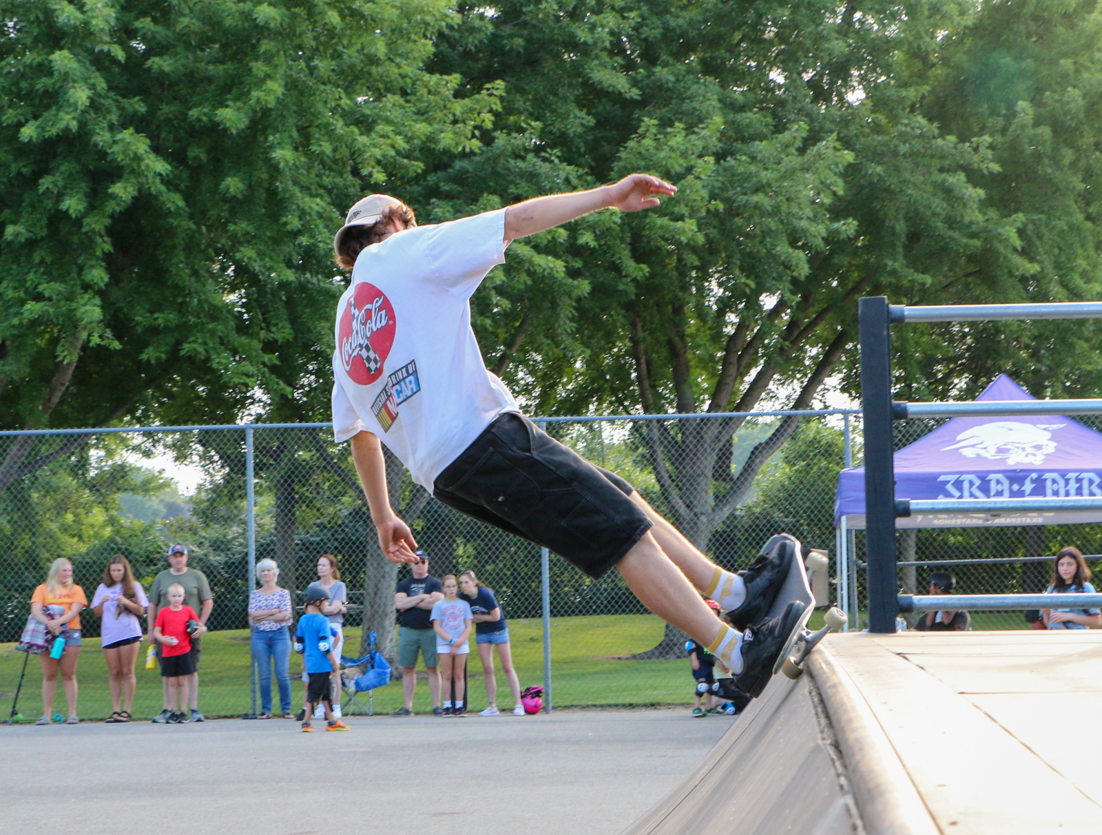 A low-angle, full shot shows a person on a skateboard leaning far back, almost parallel to the ground, with their arms outstretched for balance. They are wearing a white t-shirt with a red circular logo, black shorts, and white socks with black shoes. The skateboard is black with white wheels and is positioned on the edge of a concrete ramp.