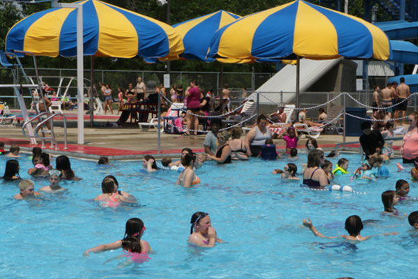 A crowded outdoor swimming pool on a sunny day, with many people, including children and adults, swimming and playing in the water. In the background, there are several large blue and yellow striped umbrellas providing shade over a paved area where more people are relaxing on lounge chairs.