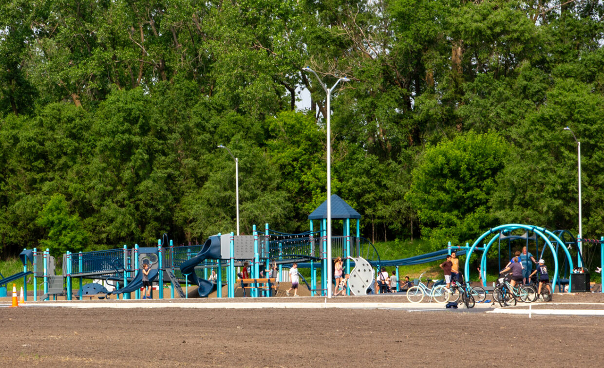 A wide, outdoor shot captures a large, modern inclusive playground with blue and gray structures, set against a backdrop of a dense green forest. The playground features multiple slides, climbing elements, and swings, including an accessible swing set on the right. Several people, including children and adults, are visible on the playground, with some enjoying the equipment and others standing nearby.