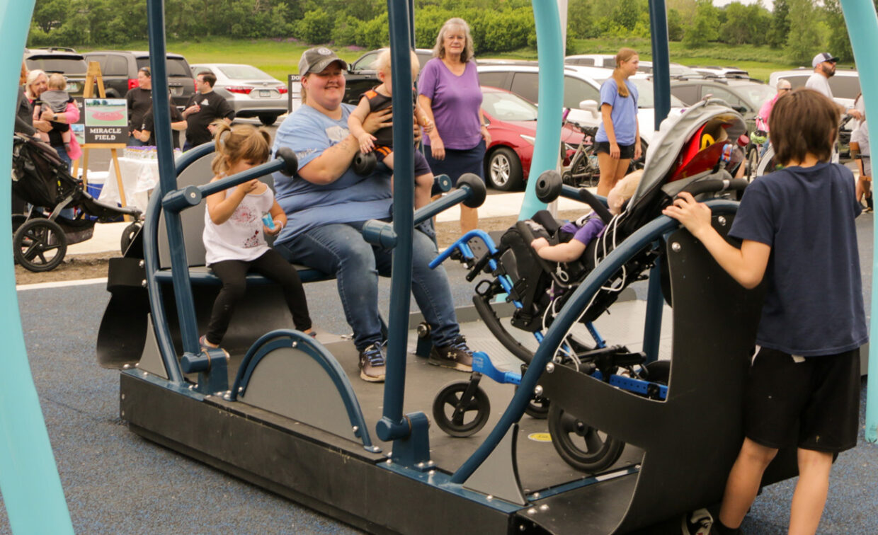A group of people, including adults and children, are gathered around a blue and black inclusive swing set on a playground. In the center, a woman in a blue shirt and jeans is seated on the swing, holding a baby, with a young girl in a white shirt and pigtails seated next to her. To the right, a child with dark hair in a dark blue shirt and black shorts stands next to a wheelchair-accessible swing, which appears to have a person seated in it.