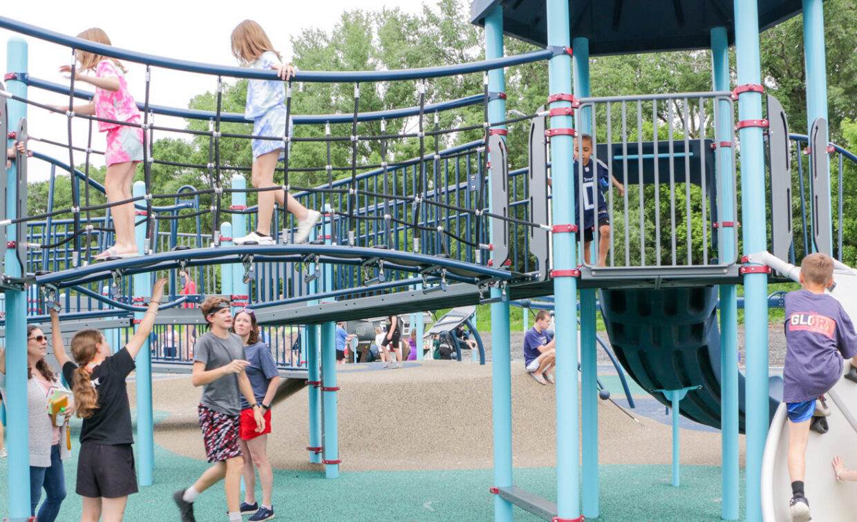 Children are actively playing on a large, modern playground structure with blue and gray elements, including a rope bridge, climbing walls, and slides.