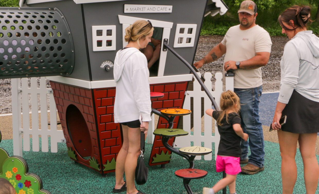 A playground scene features a "Market and Cafe" playhouse with a grey upper level, white windows, and a red brick-patterned base. A young child in pink shorts and a black shirt is ascending colorful, leaf-shaped steps that lead up to the playhouse. An adult male in a beige t-shirt and baseball cap stands near the steps, looking at the child, while two adult females, one in a white hoodie and black shorts, and another in a grey top and black skirt, stand nearby.