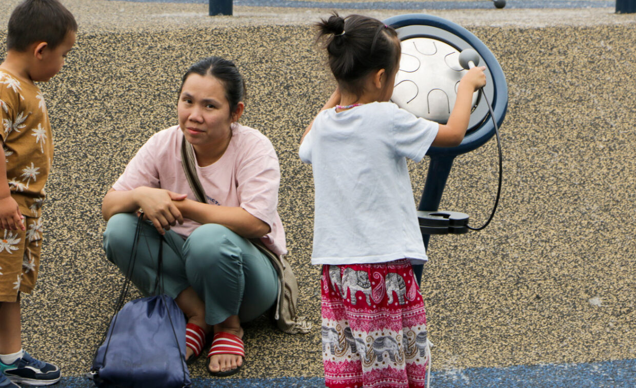 A mother squats on a speckled, light brown playground surface, looking towards the viewer. She wears a light pink t-shirt, green pants, and red sandals, with a blue backpack resting next to her. To her left, a young boy in a light brown patterned outfit stands looking at her. To her right, a young girl in a white t-shirt and red and white patterned pants stands with her back to the viewer, interacting with a blue, drum-like musical play element.