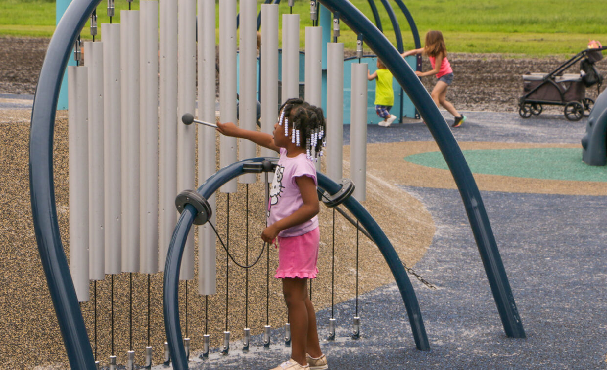 A young Black girl with braided hair and white beads, wearing a pink t-shirt and pink shorts, plays a large outdoor chimes instrument on a playground. She holds a mallet and is about to strike one of the long, silver-colored tubes. The chimes are set within a blue arched frame.