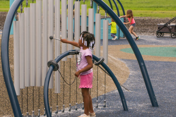 A young Black girl with braided hair and white beads, wearing a pink t-shirt and pink shorts, plays a large outdoor chimes instrument on a playground. She holds a mallet and is about to strike one of the long, silver-colored tubes. The chimes are set within a blue arched frame.