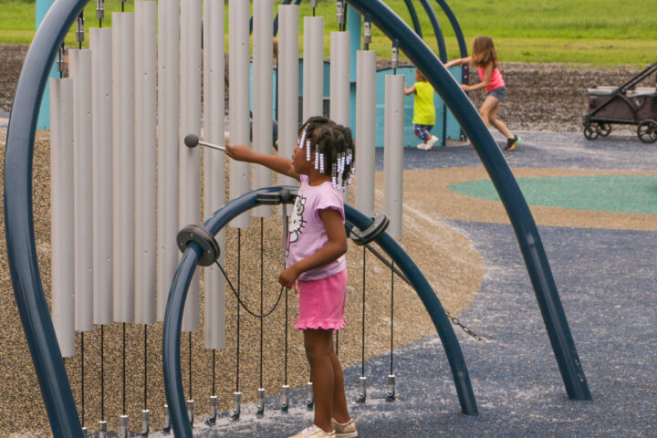 A young Black girl with braided hair and white beads, wearing a pink t-shirt and pink shorts, plays a large outdoor chimes instrument on a playground. She holds a mallet and is about to strike one of the long, silver-colored tubes. The chimes are set within a blue arched frame.
