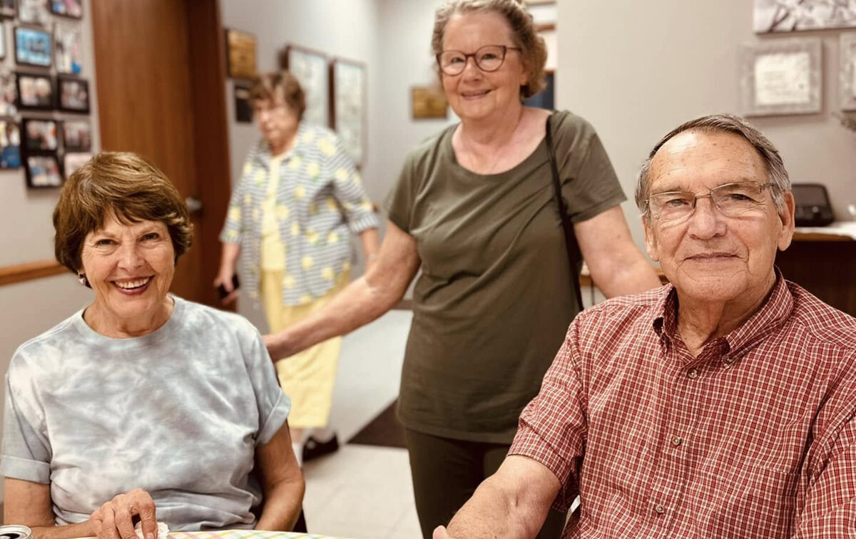 Query successful Three seniors are in what appears to be a communal or dining area. In the foreground on the left, a woman with short brown hair and a tie-dye shirt smiles at the camera. To her right, a man with glasses and a red plaid shirt also smiles, looking towards the camera. Standing behind them, a woman with glasses and an olive green shirt smiles and looks towards the camera. In the background, another person in a light-colored top is partially visible
