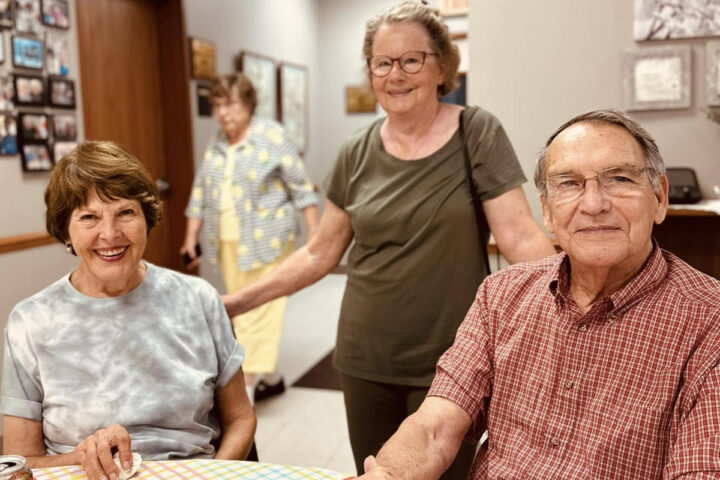 Query successful Three seniors are in what appears to be a communal or dining area. In the foreground on the left, a woman with short brown hair and a tie-dye shirt smiles at the camera. To her right, a man with glasses and a red plaid shirt also smiles, looking towards the camera. Standing behind them, a woman with glasses and an olive green shirt smiles and looks towards the camera. In the background, another person in a light-colored top is partially visible