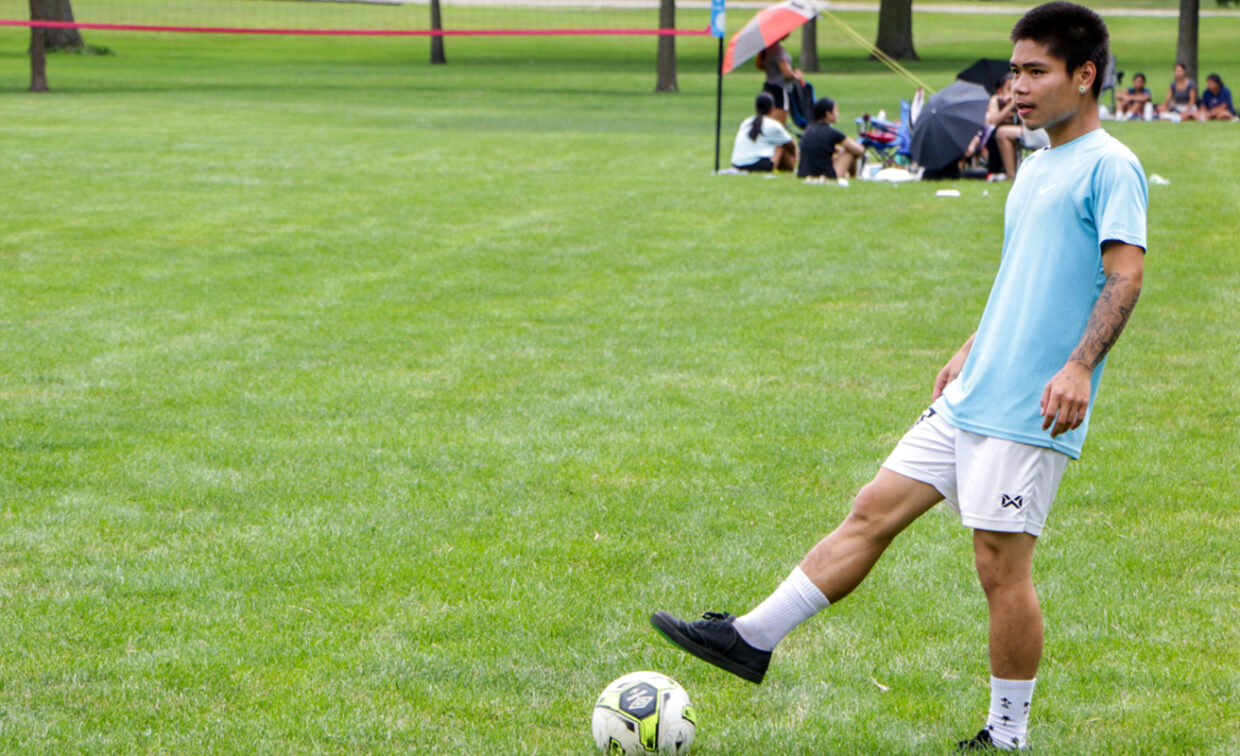 A young man in a light blue t-shirt and white shorts is playing soccer on a green field. He is in the process of kicking a black and white soccer ball with his right foot. He wears black athletic shoes and white socks. In the background, other people are visible on the grass, some sitting under umbrellas, suggesting spectators or other participants in a park setting.