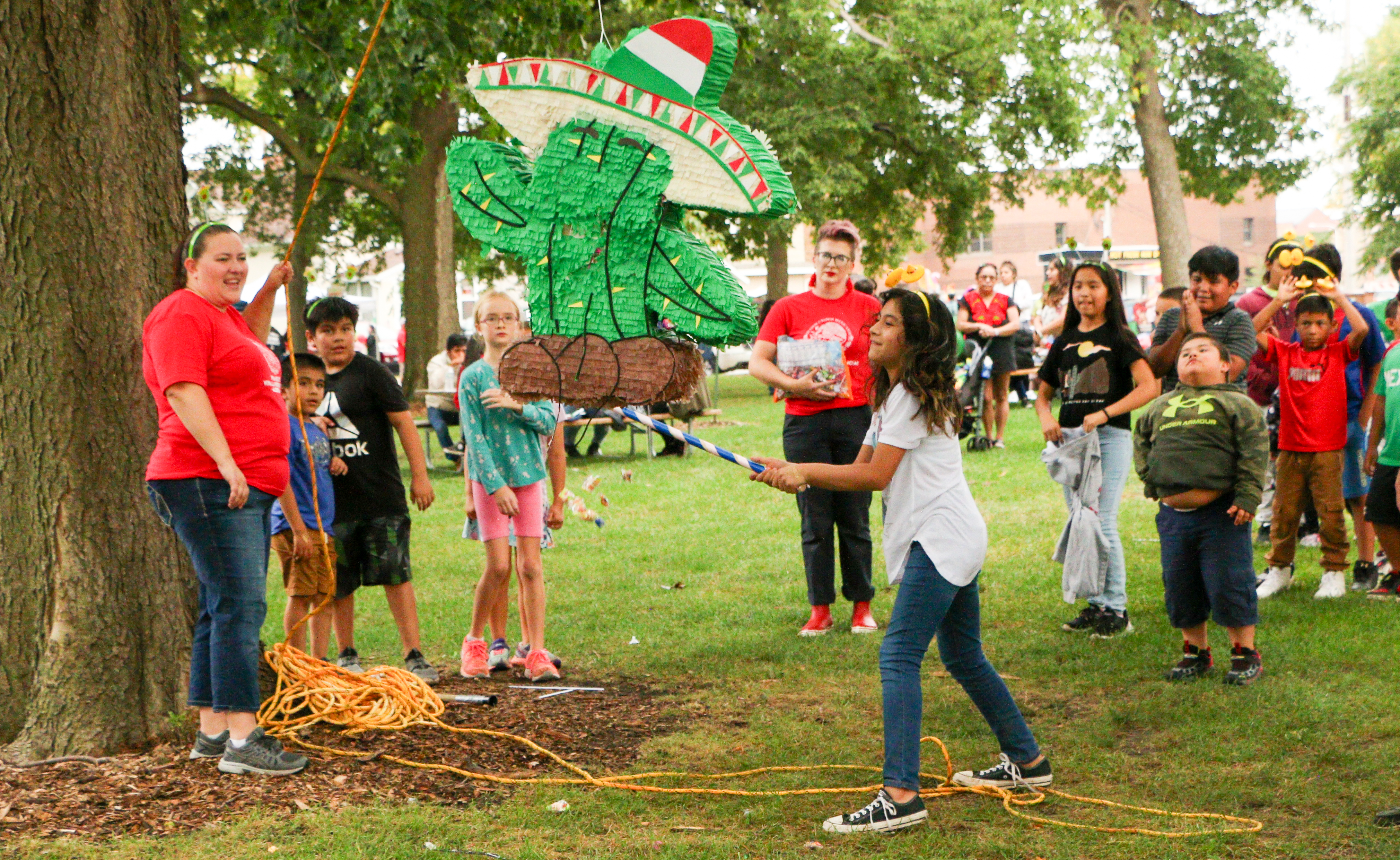 A group of children and adults are gathered around a piñata shaped like a green cactus wearing a red, white, and green sombrero, hanging from a tree branch in a grassy park. A young girl in a white shirt and blue jeans, wearing a yellow headband, swings a decorated stick at the piñata.