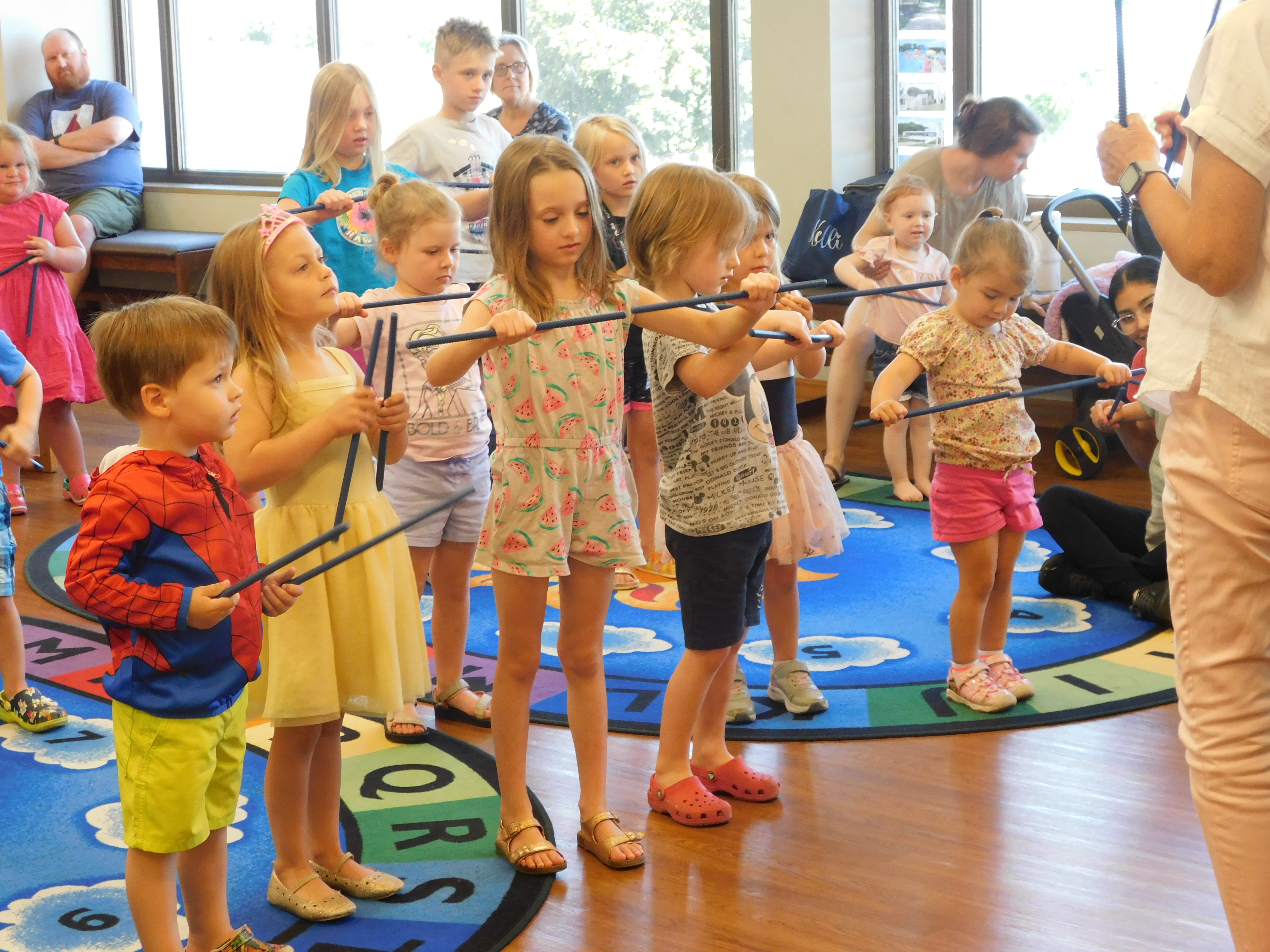 A group of young children stands on a colorful blue rug with alphabet letters, holding long, thin sticks. They appear to be participating in a music or movement activity. Several adults are visible in the background, observing the children. The room has large windows, letting in natural light.