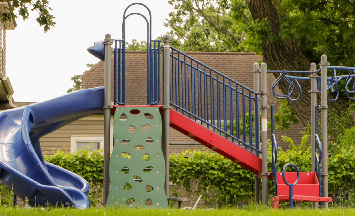 A brightly colored playground set stands on a patch of green grass. The main structure features a curved blue slide on the left, a green climbing wall with various shaped cutouts in the center, and a red staircase with blue railings leading up to a platform. To the right, there's a blue monkey bar section with red swinging rings and a small red swing seat.
