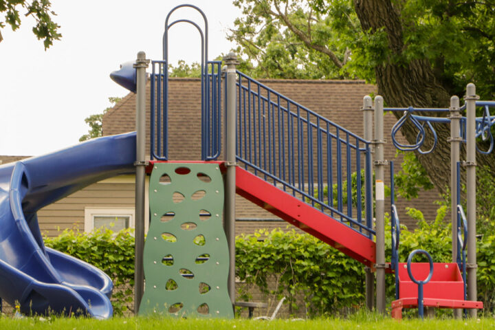 A brightly colored playground set stands on a patch of green grass. The main structure features a curved blue slide on the left, a green climbing wall with various shaped cutouts in the center, and a red staircase with blue railings leading up to a platform. To the right, there's a blue monkey bar section with red swinging rings and a small red swing seat.