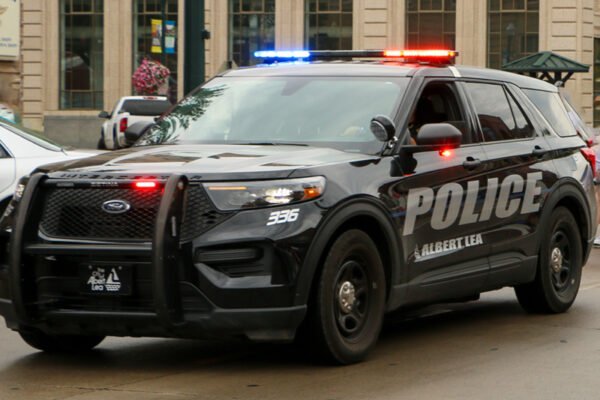 A black Albert Lea Police Ford Interceptor SUV with red and blue lights flashing, on a city street.