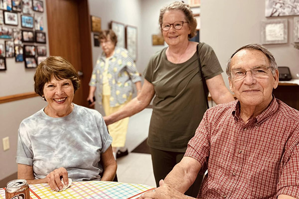 A group of four seniors, two men and two women, are gathered around a table indoors. In the foreground, a woman with short brown hair and a tie-dye shirt smiles at the camera, holding a tissue. To her right, a man in a red and white plaid shirt also smiles at the camera. In the background, another woman in a green shirt stands behind them, looking forward with a slight smile. Further back and out of focus, a fourth woman in a yellow and white checkered shirt is partially visible.