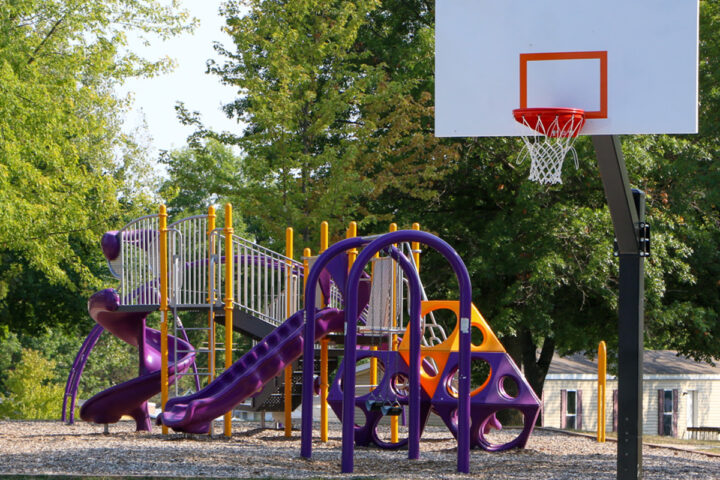 A purple and yellow playground set with slides and climbing structures stands next to a basketball hoop in a park with green trees and some houses in the background.