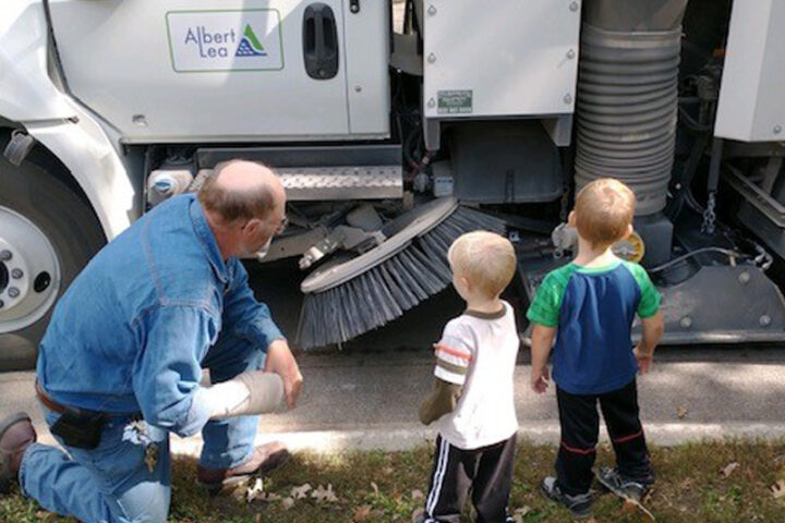 A city worker in a denim jacket and jeans kneels next to a street sweeper, pointing at its large rotating brush. Two young children, seen from behind, look on with interest. The side of the white street sweeper has a green logo that says 