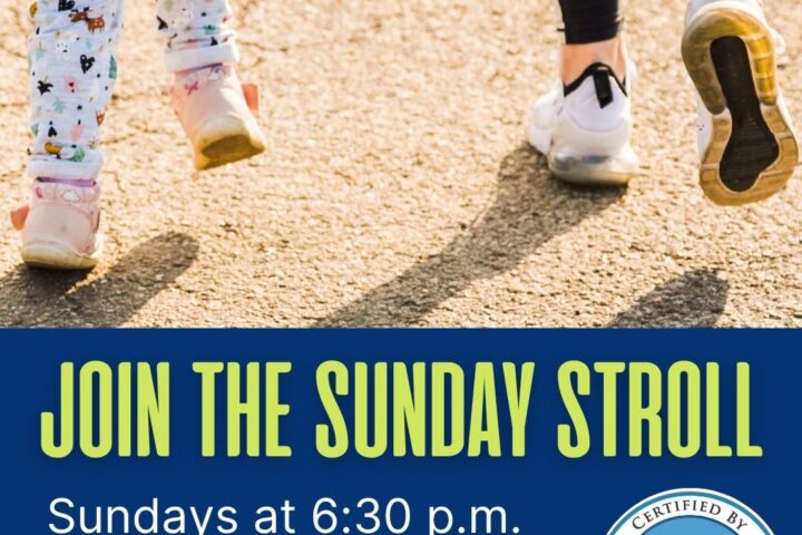 An image shows an adult and a young child walking away from the camera, their legs and feet visible. The adult is on the right, wearing dark pants and white sneakers with blue accents. The child is on the left, wearing light-colored pants with patterns and pink shoes. The ground is a light-colored asphalt or paved surface, with sunlight creating shadows. Below them, a dark blue banner reads 