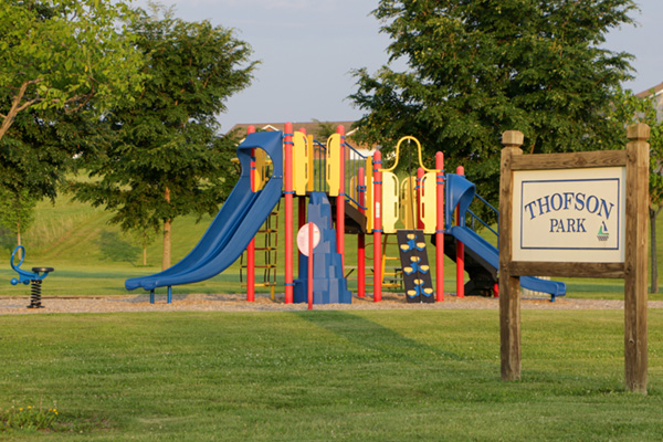 A bright, multi-colored playground with blue slides, red poles, and yellow platforms sits on a wood-chip base in the middle of a green grassy park. A wooden sign on the right says 