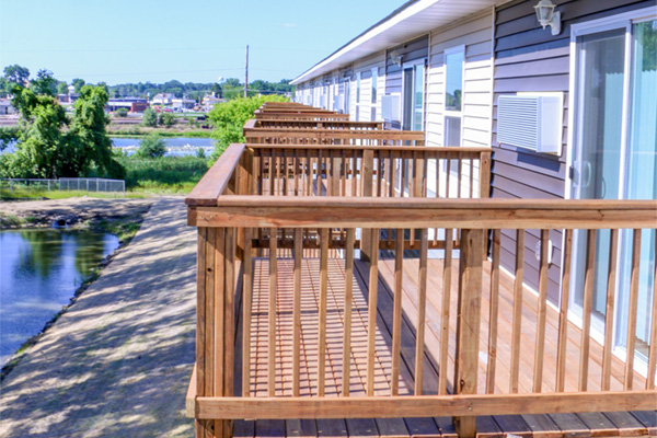 A row of apartments with individual wooden decks overlooks a body of water and a grassy area. The apartment building is two stories high, with light grey siding on the upper level and a darker grey siding below. Each apartment has a sliding glass door leading to a private deck with a wooden railing. Some apartments also have air conditioning units mounted below the windows. In the background, across the water, there are trees and some buildings, possibly part of a town or industrial area. The sky is blue with a few clouds.