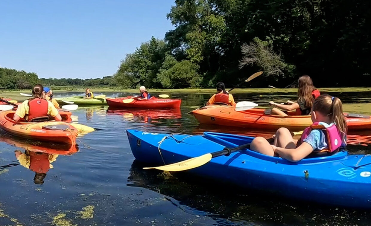A group of adults and children in colorful kayaks paddle on a calm, tree-lined waterway under a clear sky.
