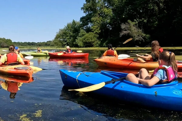 A group of adults and children in colorful kayaks paddle on a calm, tree-lined waterway under a clear sky.